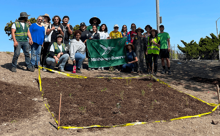 Group of volunteers pose on a sunny hillside behind a mulched, newly planted garden bed bordered with yellow caution tape, holding a “Mini Nature Reserve” banner.