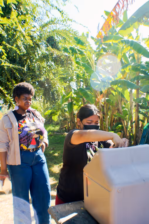 Two participants at a garden workshop; one operates equipment under banana trees in bright sunlight.
