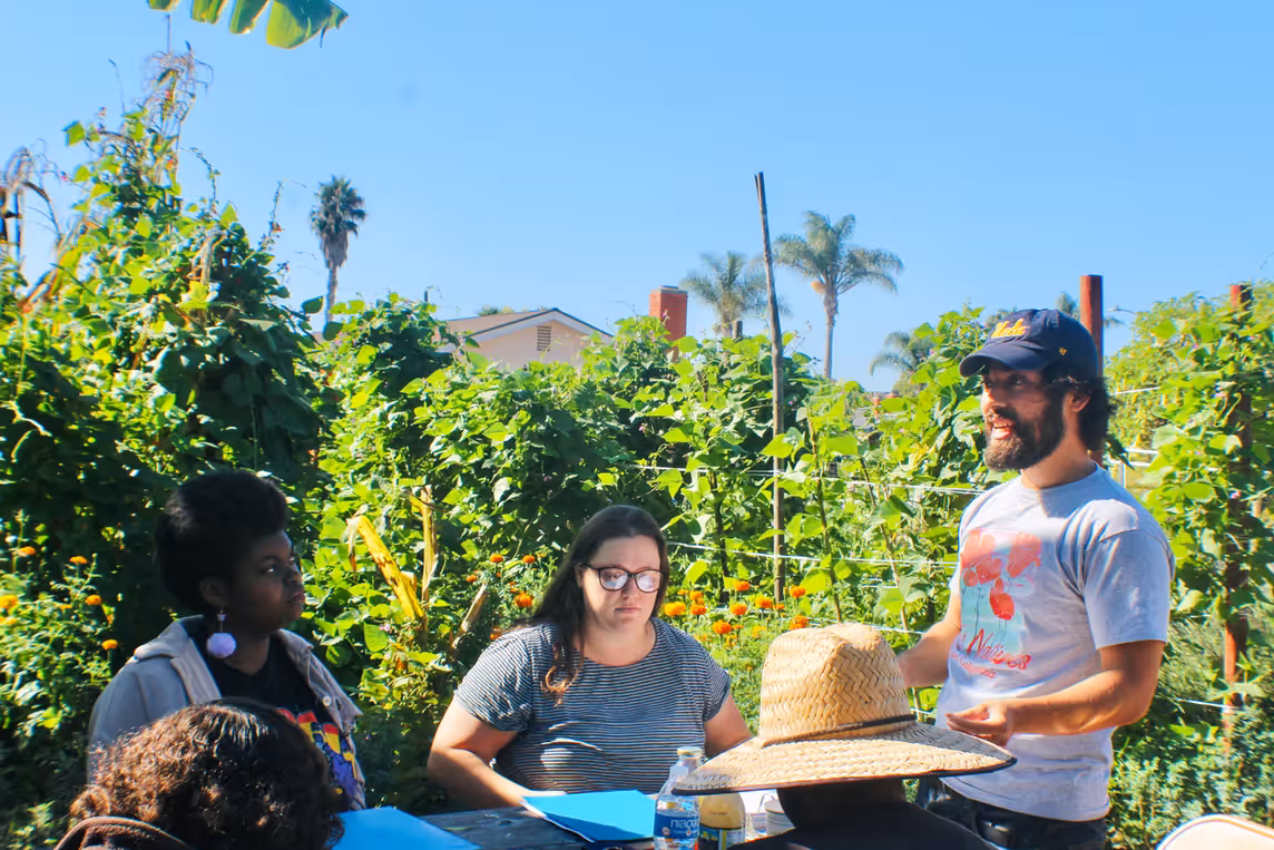 A small group gathers in a lush community garden, listening to a speaker in a cap and t-shirt leading an outdoor workshop surrounded by vines and palm trees.